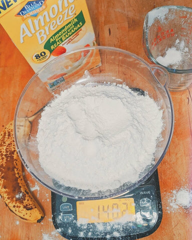 Truweigh Wave scale has a large bowl of flour on it, with flour spilled outside of the bowl. The scale is on an orange background, and a measuring bup, banana and almond milk are also in the frame.