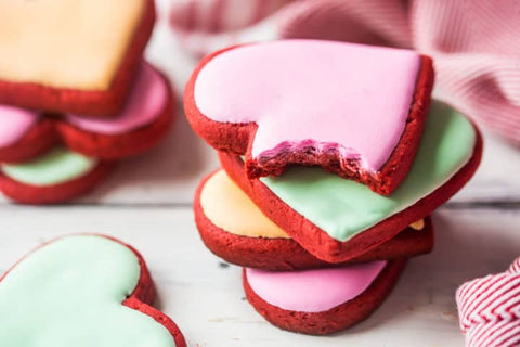 A few stacks of the red velvet heart-shaped Valentine's Day cookies are shown. The front stack has a cookie with pink frosting on top with a bite taken out of it. Photo credit to the Baking a Moment blog.