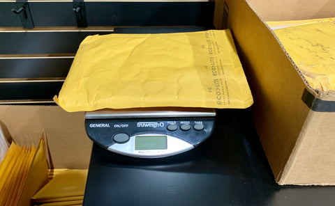 The Truweigh General bench scale is weighing a yellow padded envelope that is being prepared for shipment.