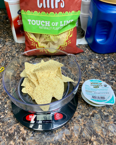 The Truweigh Vortex kitchen scale sits on a granite counter. It has a serving of lime tortilla chips being weighed, and an individual guacamole pack is next to the scale with the seal still in tact. The bag of chips is behind the scale.