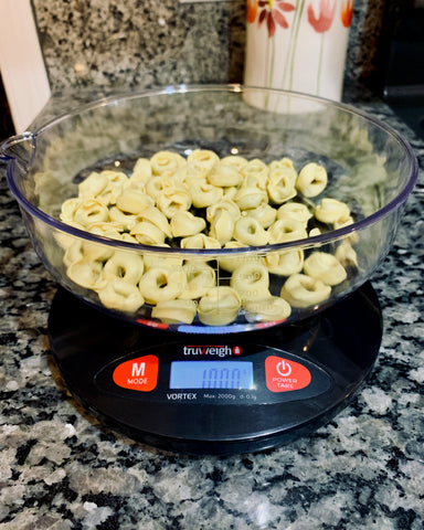 The Truweigh Vortex digital kitchen scale sits on a granite countertop and the weighing bowl is filled with dry tortellini pasta.