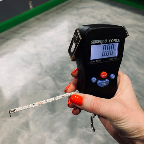 A female hand with red nail polish holds the Truweigh Force Digital Hanging scale indoors with the tape measure extended, and held between her thumb and middle finger.
