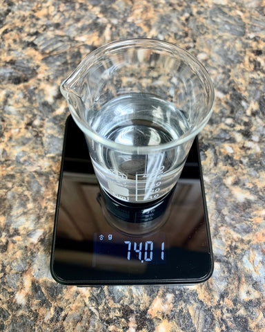 A small beaker of water is sitting on top of a Truweigh Storm digital scale. The scale is all black with gray numbers reading the weight 74.01 grams. The scale is on a Formica counter top