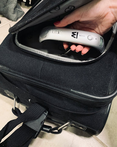 A female hand holds the Truweigh Hook digital hanging scale inside the front pocket of a black carry-on suitcase. The suitcase is laying on its side on the ground.