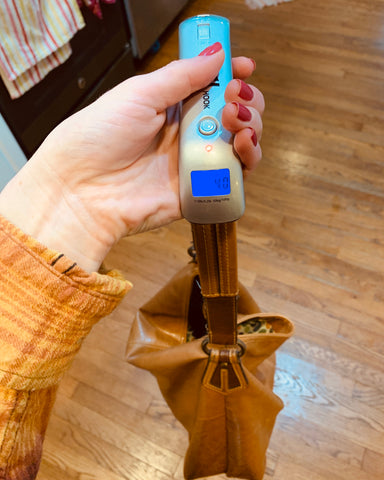 A female hand with pink nail polish and a mustard yellow sleeve hold the Truweigh Hook digital hanging scale. The scale is weighing a brown leather purse.