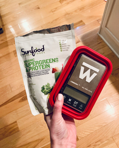 The Truweigh Crimson digital food scale is turned off, cover on inside the collapsed silicone weighing bowl. A female hand holds the scale and a packet of superfood protein powder in her hand above a wood floor.
