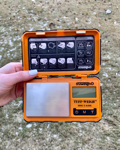 A white female hand holds the orange Truweigh Tuff-Weigh digital scale with the cover open showing the entire scale. The scale is held outside against a grassy background.