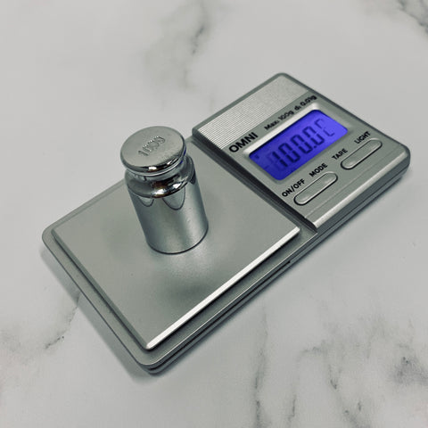 The Truweigh Omni digital scale is being calibrated with a 100g official calibration weight. The screen is on and reads 100g, and the scale sits on a white marble counter.