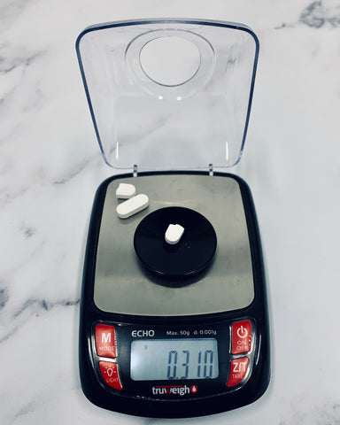 The Truweigh Echo Milligram digital scale sits on a white marble background and is weighing half of a white pill. The other half and one other full pill are sitting in the left top corner of the scale.