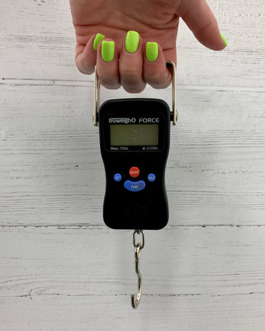 A female hand with bright green nail polish holds the Truweigh Force Digital Hanging scale in front o f a white wooden background. The scale is held by her second, middle and ring fingers.