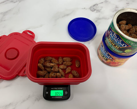 The Truweigh Crimson digital food scale is weighing a portion of almonds in the collapsible silicone bowl. Two cans of almonds are stacked to the right, the top can is open with the lid sitting next to the cans. Everything sits on a white marble surface.