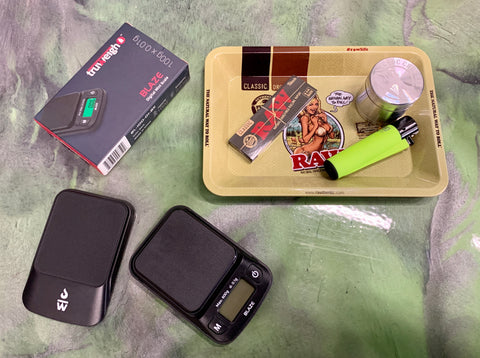 The Truweigh Blaze digital mini scale is set out on a counter next to its box and the cover is set to the side. It sits next to a small RAW rolling trays with RAW rolling papers and a yellow Clipper lighter.