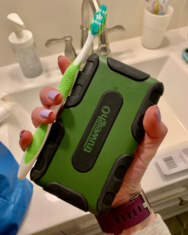 A female hand holds the green Truweigh Tuff-Weigh with a green and white toothbrush in front of a sink, preparing to clean the scale.