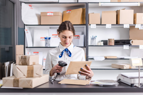 Young woman at post office counter scanning in mail and packages to be sent out. She stands in front of a bookcase filled with other mail and packages