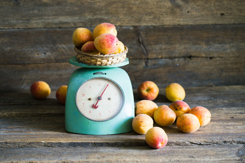 A light teal spring balance scale holds several peaches with a pile of peaches sitting next to it. The scale and fruits sit on a rustic wood counter top in front of the same wood background.
