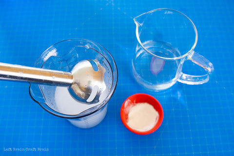 The ingredients for the edible bubble experiment sit together on a blue counter. There is a measuring cup with electric mixer in it with the sodium alginate mixture, a small bowl with powder, and a pitcher of water. Credit to Left Brain Craft Brain