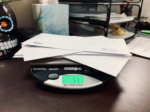 Truweigh general compact bench scale with a pile of letters being weighed. The scale sits on a dark wood desk with other office supplies in the background.