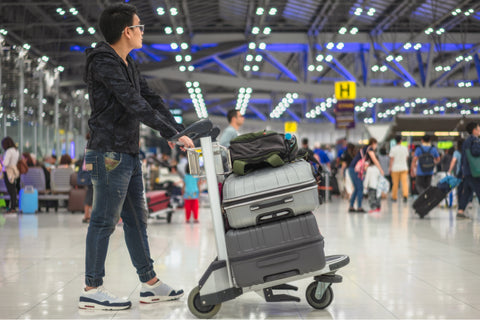 A young Asian man pushes a baggae trolley carrying two large pieces of luggage and a backpack. He is walking through a fairly busy airport.