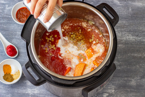 A hand pours a white liquid from a can into an Insta pot pressure cooker filled with sweet potato curry. Other ingredients sit in small bowls and a spoon on the gray counter.