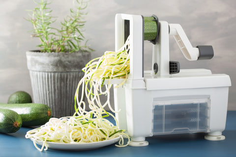A vegetable spiralizer is being used on a zucchini with a plant in the background and several untouched zucchinis in frame. There is a pile of zucchini noodles on a plate beneath the machine.
