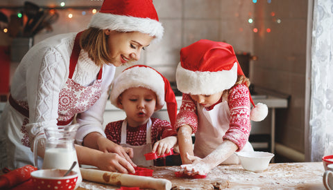 A mom and two young kits are making Christmas sugar cookies wearing Santa hats. The little girl is using a cookie cutter to cut out a shape, while the mom and little boy also hold cookie cutters and watch the little girl.