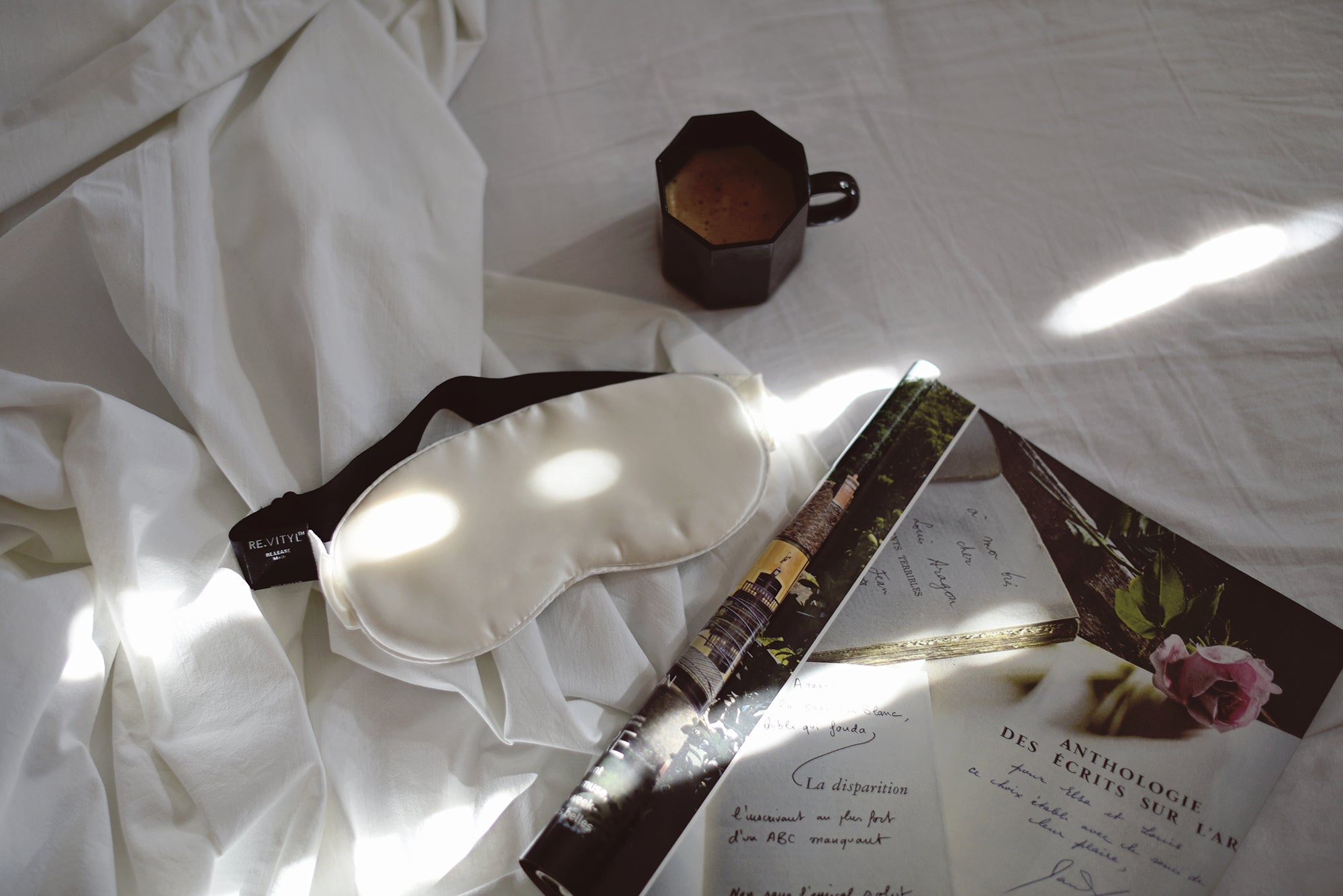 White silk sleep mask with crystals on a bed by a cup of coffee and a magazine