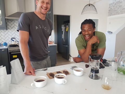 Philip Snyman and Cecil Afrika standing at the kitchen counter when we did our first 18 coffee blend cupping, they are both laughing out of the surprise that our coffee dream might soon become a reality. There is bag of coffee, coffee grinder (Handground kickstarter project) and some cups on the counter.