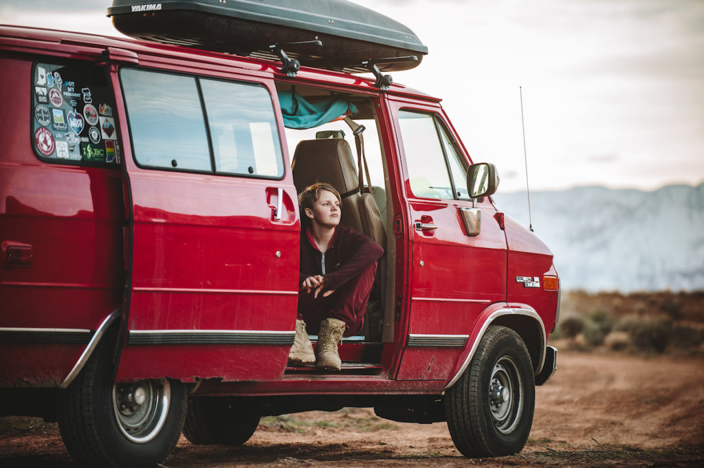A red van with the door open and a teen sitting in the doorway gazing into the distance.