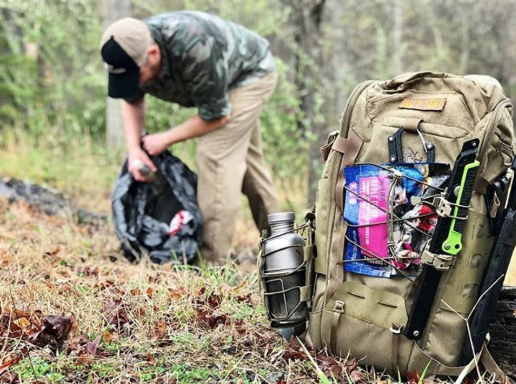 A pouch of apple crisp is strapped to the outside of a tactical backpack, with someone picking up litter in the background