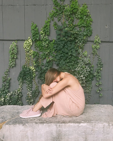 Woman in pale pink outfit covering face with hair and sitting in front of a wall covered in English ivy leaves, for Ivy Leaf Skincare