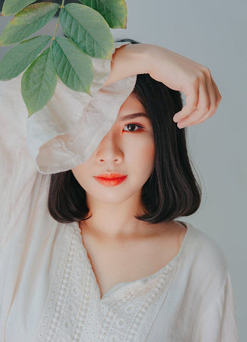 A woman in white standing behind a green tree branch, covering half of her face with her arm, for Ivy Leaf Skincare