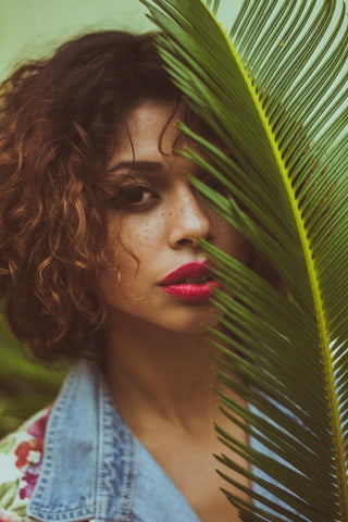 A woman standing behind a palm tree leaf, covering half of her face, for Ivy Leaf Skincare