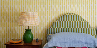fern mustard wallpaper set in a single bedroom a headboard upholstered in luna turmeric hand block printed fabric all designed by molly mahon finished off with a pleated fabric lampshade in seed mustard sitting on top of a honey pot lamp base