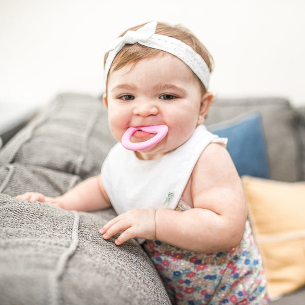 A cute infant girl playfully smiling and chewing one a pink first teether made from silicone while standing on a couch and pillows.