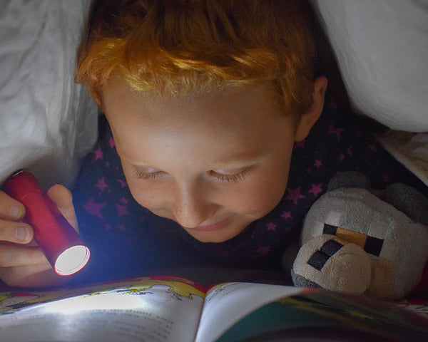 Little kid reading a book with a flashlight under a blanket