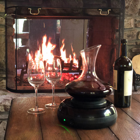 Aerisis wine aerator with 2 wine glasses and bottle of red wine on a wooden coffee table with fireplace in the background