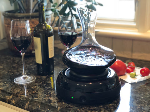 Aerisi wine aerator with decanter with red wine sitting on dark granite counter top near a kitchen window with 2 glasses of red wine and a wine bottle and green plant behind it