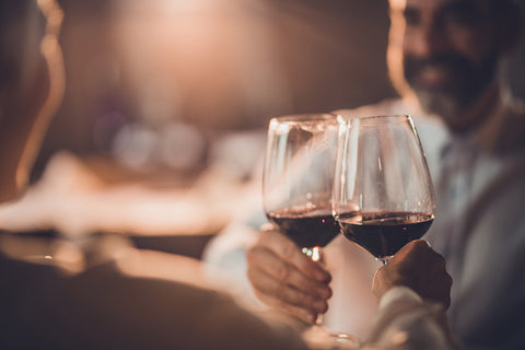 Gentleman in a white dress shirt and beard sitting at a restaurant table facing us, his companion sitting across from him with their back toward us, toasting each other each holding a glass of red wine, soft warm light in the background