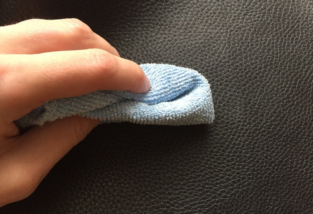 a close up of black vegan leather. There is a hand in view holding a small blue terrycloth and is gently scrubbing the item clean. 