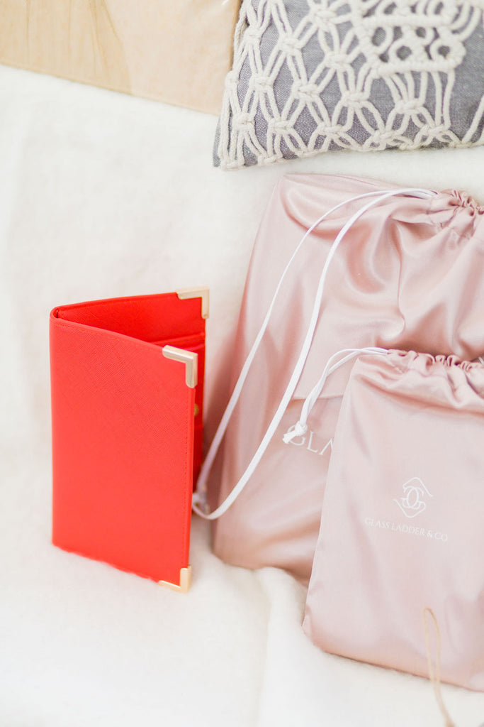 A red Amelia wallet is on a white fluffy, background. It is standing upright and is partially opened. The gold corner bumpers are shiny against the bright red material. To the right of the Amelia is a small blush pink dust bag, partially in view. 