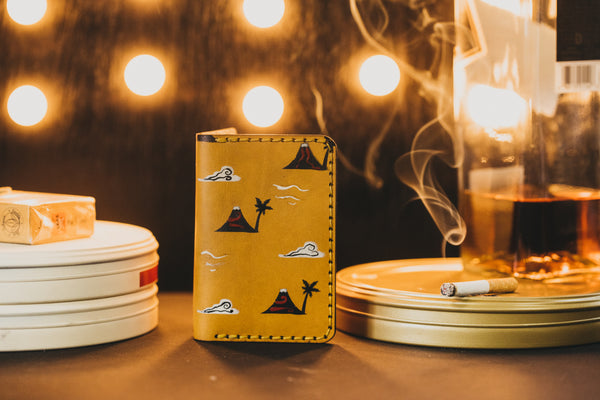 A Yellow wallet with clouds sits on a table with film reels on both sides of wallet. One reel has a cigarette and whiskey.