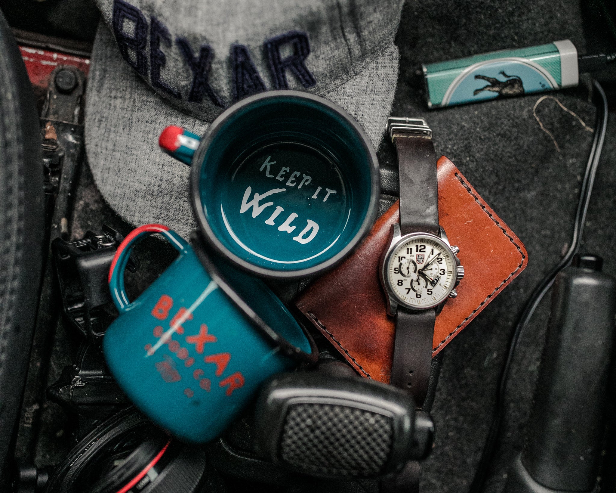Close up shot of a gray heather Ebbets x Bexar Cap slightly out of focus. A Blue enamel coffee mug with red accents sits on top with visible text on the bottom inside of mug. Keep it Wild 