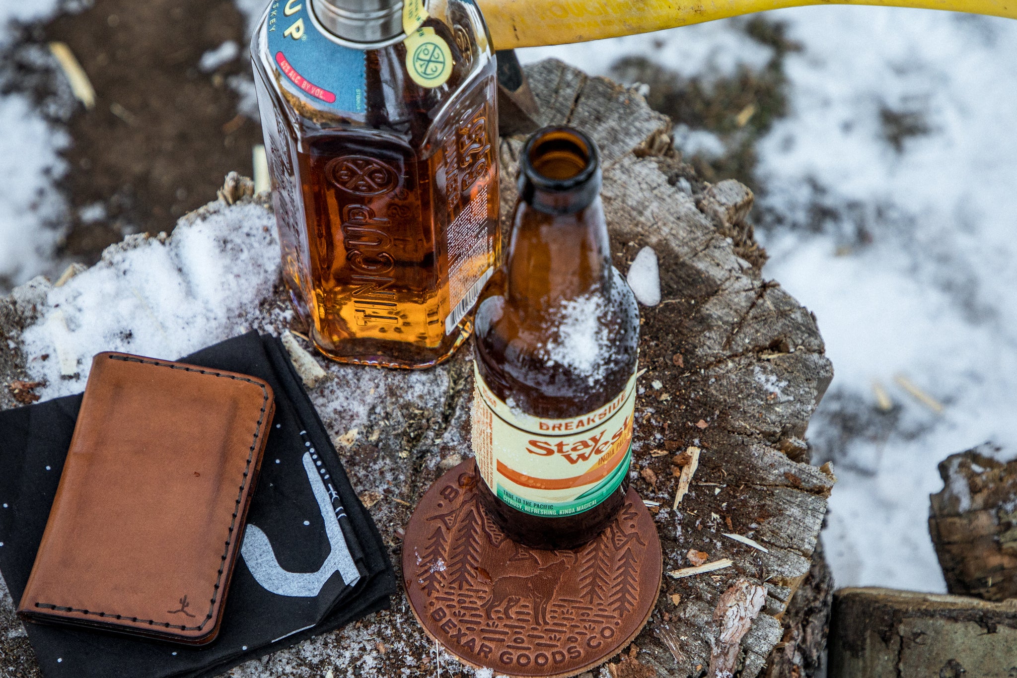 A cold and snowy stump with a Black Bandana, Bexar goods wallet, Whiskey Bottle , Beer bottle and leather coaster sit on top of tree stump