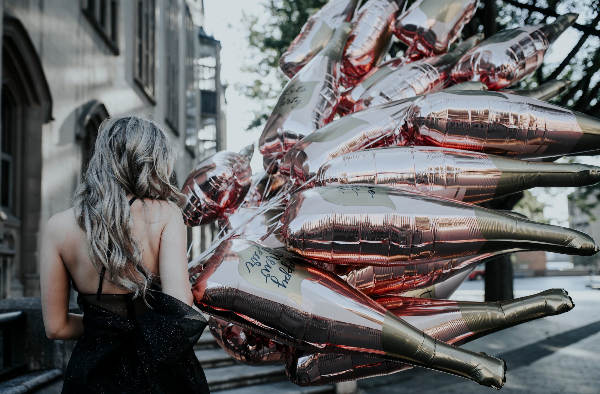 A women in a black dress walking outside holding a dozen 36-inch rose gold champagne bottle balloons with gold on the top