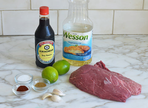 Photo of the following ingredients: raw flat iron steak, soy sauce, limes, vegetable oil, raw cloves of garlic, sugar, cumin, chili powder. 