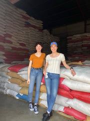 Two women smiling and leaning on bags of parchment coffee to be purchased and further processed