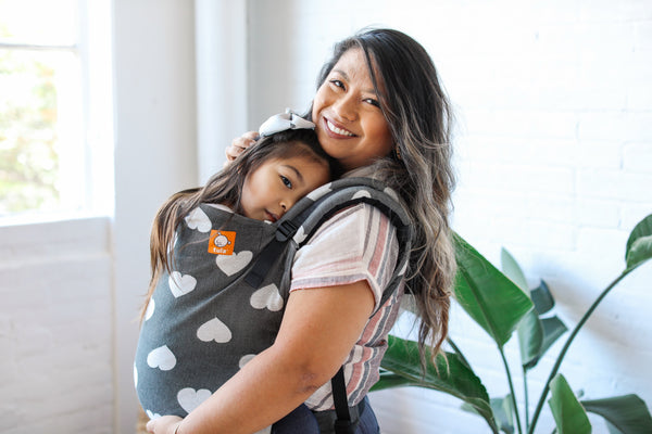A mother smiling into the camera while her Preschooler is snuggling onto her.
