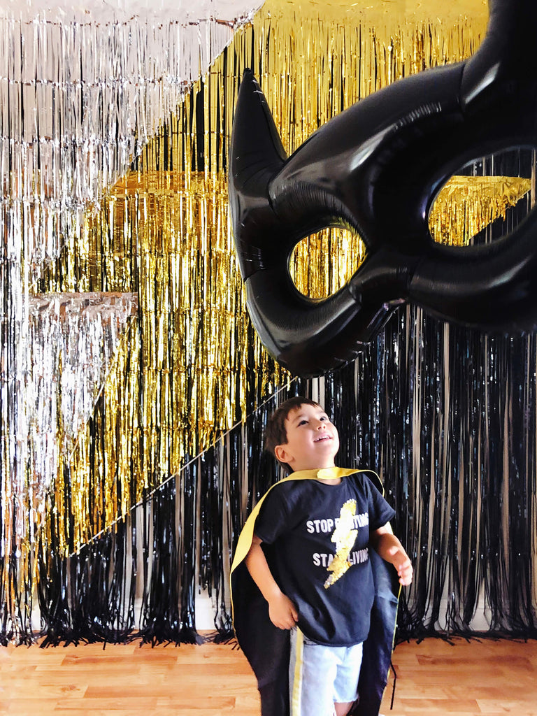 little superhero boy with black and yellow cape, looking at a giant black bat mask foil balloon in front of a black gold sliver fringe curtains made lighting bolt icon backdrop