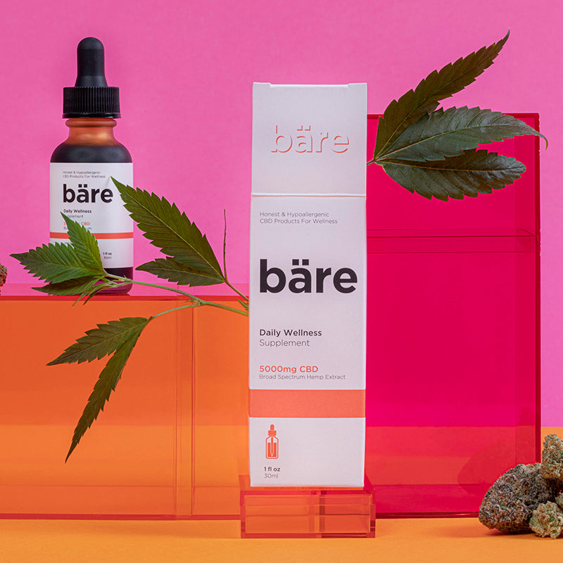 Kush Queen Bare CBD Tincture sitting on orange and pink boxes alongside cannabis plant leaves and nuggs.