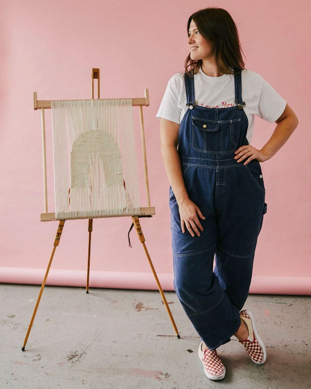 Rachel Wood wearing denim overalls and a white t-shirt posed next to her woven artwork